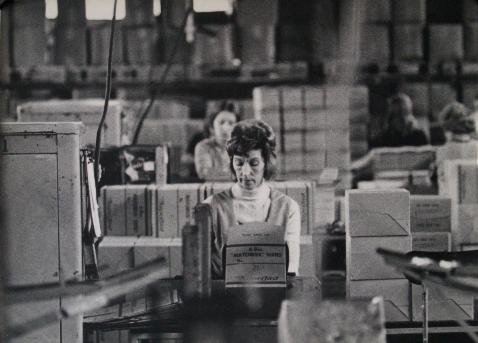 Image from ‘Women and Work’ 1975 ©Hackney Flashers. A black and white photo of a busy factory scene. Many boxes are piled up in front of and along with machinery surround women seated at tables looking down focussed on their work. One woman in the foreground and centre is the focus of the image, her concentration is clear from her expression.