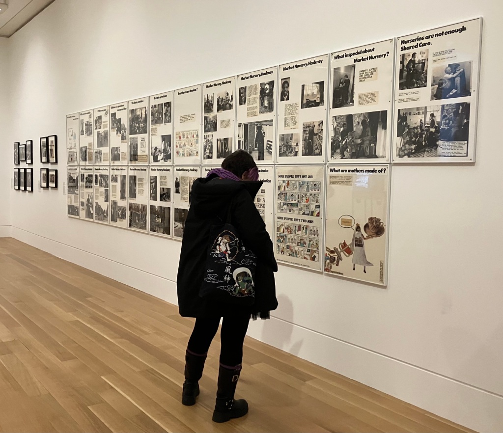 An art gallery interior (Tate Britain). Panels of Who's Holding the Baby by the Hackney Flashers is displayed on the wall. A person is looking closely at one of the panels.